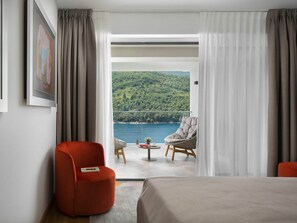 Une chambre d’hôtel avec un lit, un fauteuil rouge, une petite table ronde ornée d’une plante, et une vue sur un lac et une forêt, visible à travers de fines rideaux.