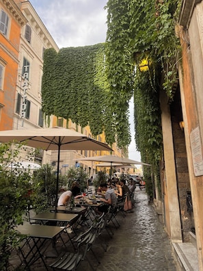 Un café en plein air, avec des tables et des chaises, entouré de bâtiments dont les murs sont recouverts de lierre.