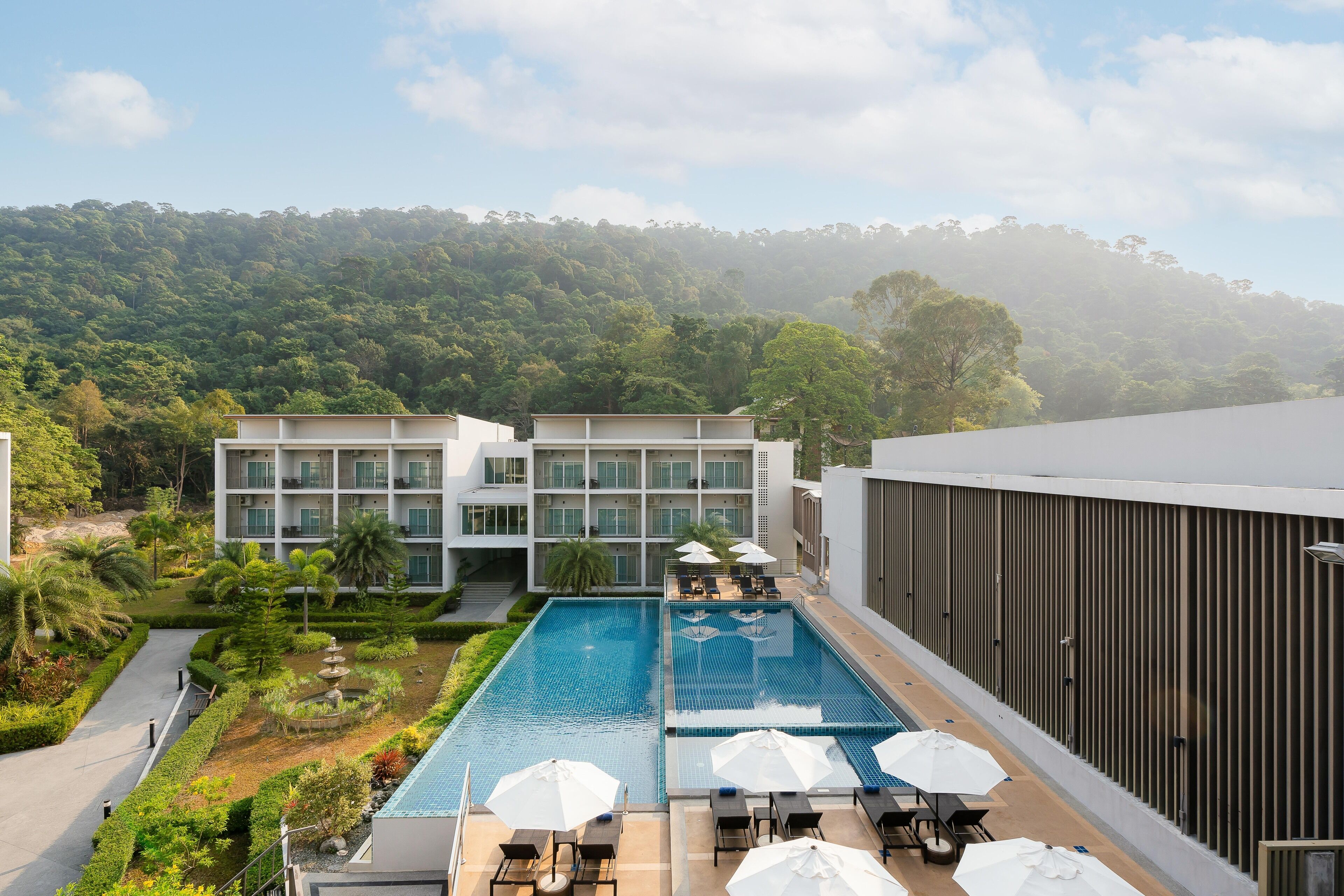 Un hotel moderno con piscina en la azotea, rodeado de exuberante vegetación y una colina boscosa al fondo.