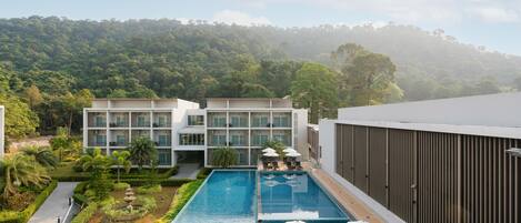 A modern hotel with a rooftop pool, surrounded by lush greenery and a forested hill in the background.
