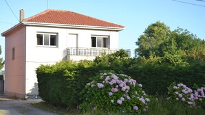 Um edifício de dois andares com telhado de telhas vermelhas, varanda com guarda-corrimão preto e uma sebe densa de flores cor-de-rosa na frente.