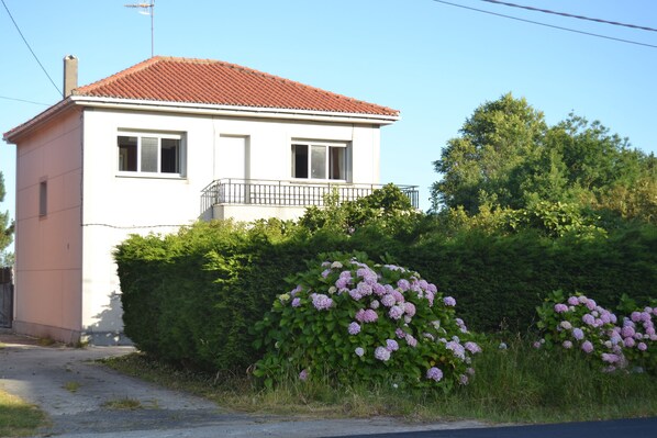 A two-story building with a red-tiled roof, a balcony with a black railing, and a dense hedge of pink flowers in front. - House with direct access to Finisterre beach (Finisterre)
