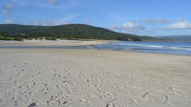 Uma extensa praia de areia com pegadas, céu claro e colinas ao longe.