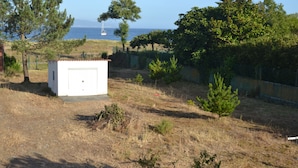 Um pequeno edifício branco com uma porta, rodeado por grama seca e vegetação rala, perto de uma cerca e árvores.