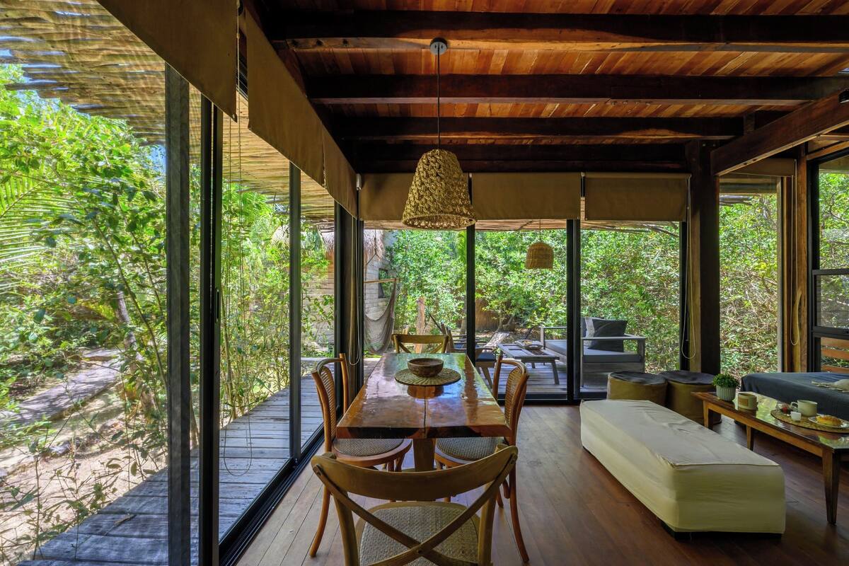 A spacious room with a wooden dining table, chairs, and a sofa. Large windows provide a view of greenery outside.