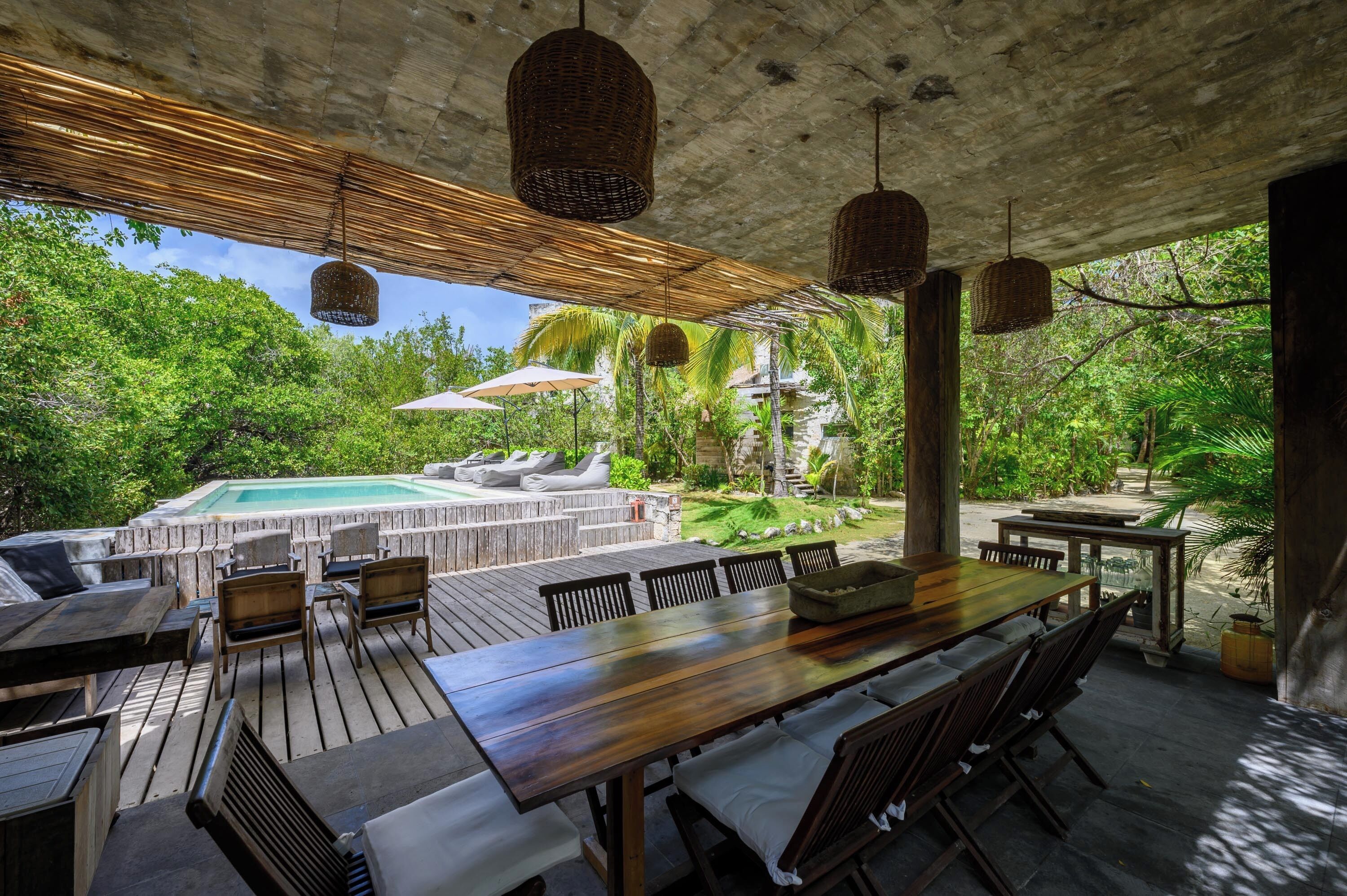 An outdoor dining area with a pool, wooden furniture, and hanging wicker lamps.