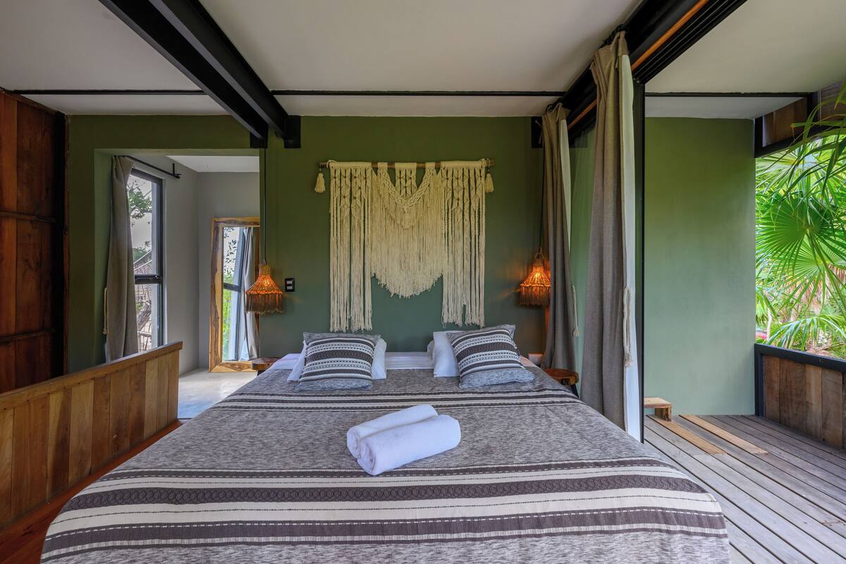 A large bed with a striped bedspread, two pillows, and a rolled towel. There is a macrame wall hanging above the bed.