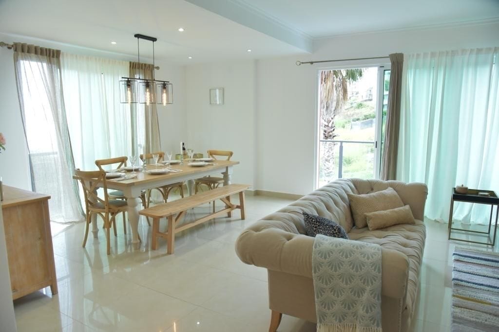 A spacious living area with a dining table, wooden chairs, a sofa, and a large window with curtains.