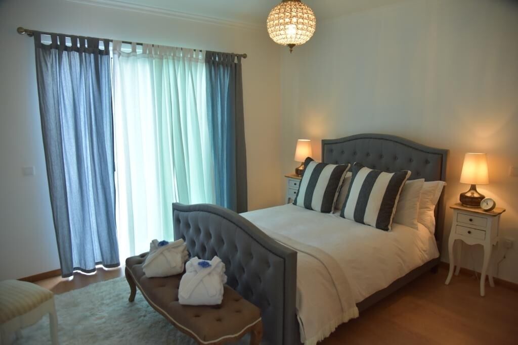 A bedroom with a large bed, two bedside tables with lamps, a bench with towels, and a chandelier.