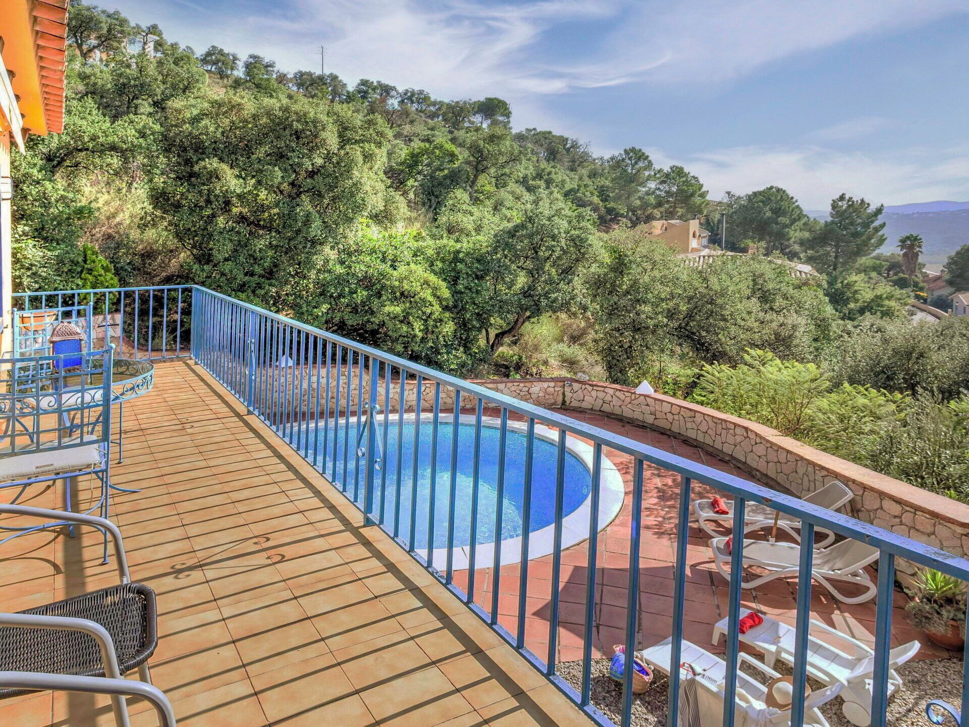A swimming pool with a wooden deck and metal railing, surrounded by greenery and overlooking a hilly landscape.