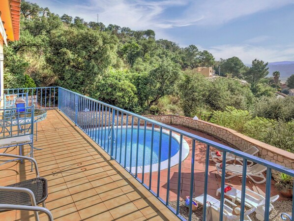 A swimming pool with a wooden deck and metal railing, surrounded by greenery and overlooking a hilly landscape.