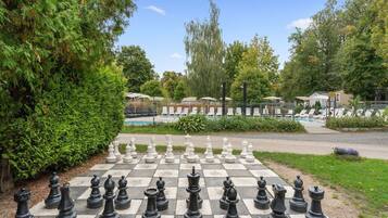 Um grande tabuleiro de xadrez ao ar livre montado em um parque, com uma piscina e espreguiçadeiras ao fundo.