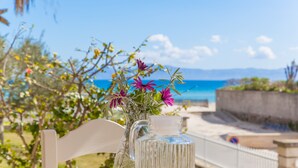 Ein Tisch mit einem weißen Stuhl und einer Karaffe mit Blumen, mit Blick auf einen Strand unter blauem Himmel, im Hintergrund ist ein Zaun zu sehen.