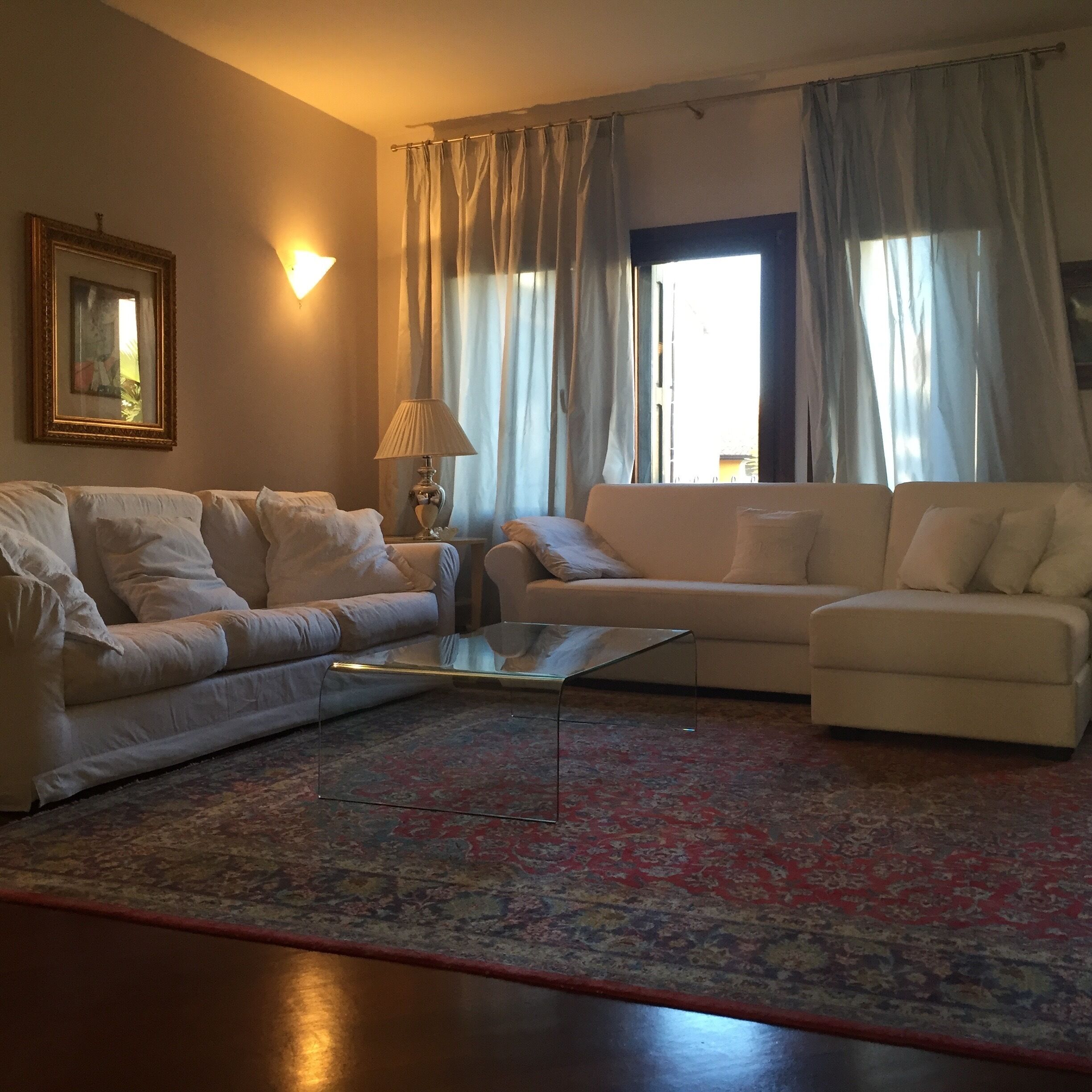Sala de estar com dois sofás brancos, uma mesa de centro de vidro, um abajur e um tapete estampado.