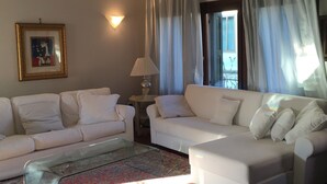 A living room with two white sofas, a glass coffee table, a lamp, a framed picture on the wall, and a view through the window.