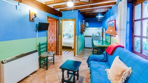 Een gezellige kamer met een blauwe bank, een kleine tafel, een groene stoel, een plafondventilator en een keukenruimte zichtbaar door een geopende deur.