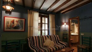 Een gezellige kamer met een gestreept bankje, een klein tafeltje en een stoel. Er is een raam met gordijnen, een ingelijste foto aan de muur en een lamp.