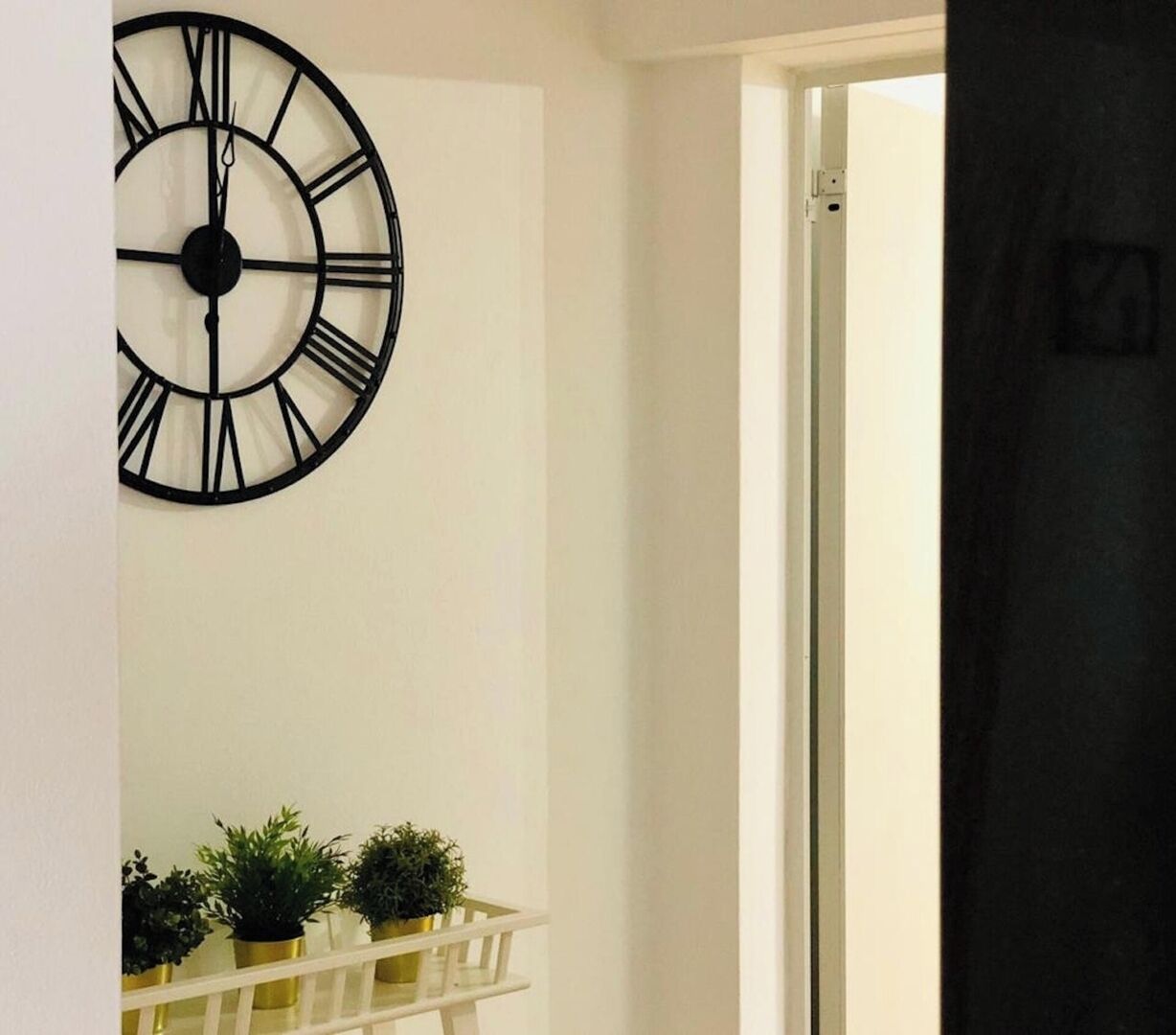Ein Zimmer mit einer großen schwarzen Uhr an der Wand, einer weißen Tür und einem kleinen weißen Regal mit Topfpflanzen.