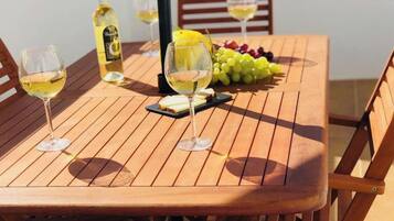 A wooden table with two glasses of white wine, a bottle of wine, a plate of grapes, and a small cheese board.
