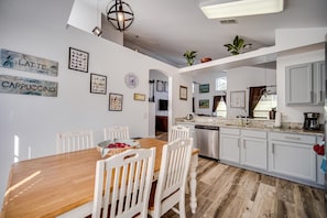 Una cocina moderna con armarios blancos, una mesa de comedor de madera y sillas. En las paredes hay cuadros enmarcados, un reloj y una planta en la encimera.