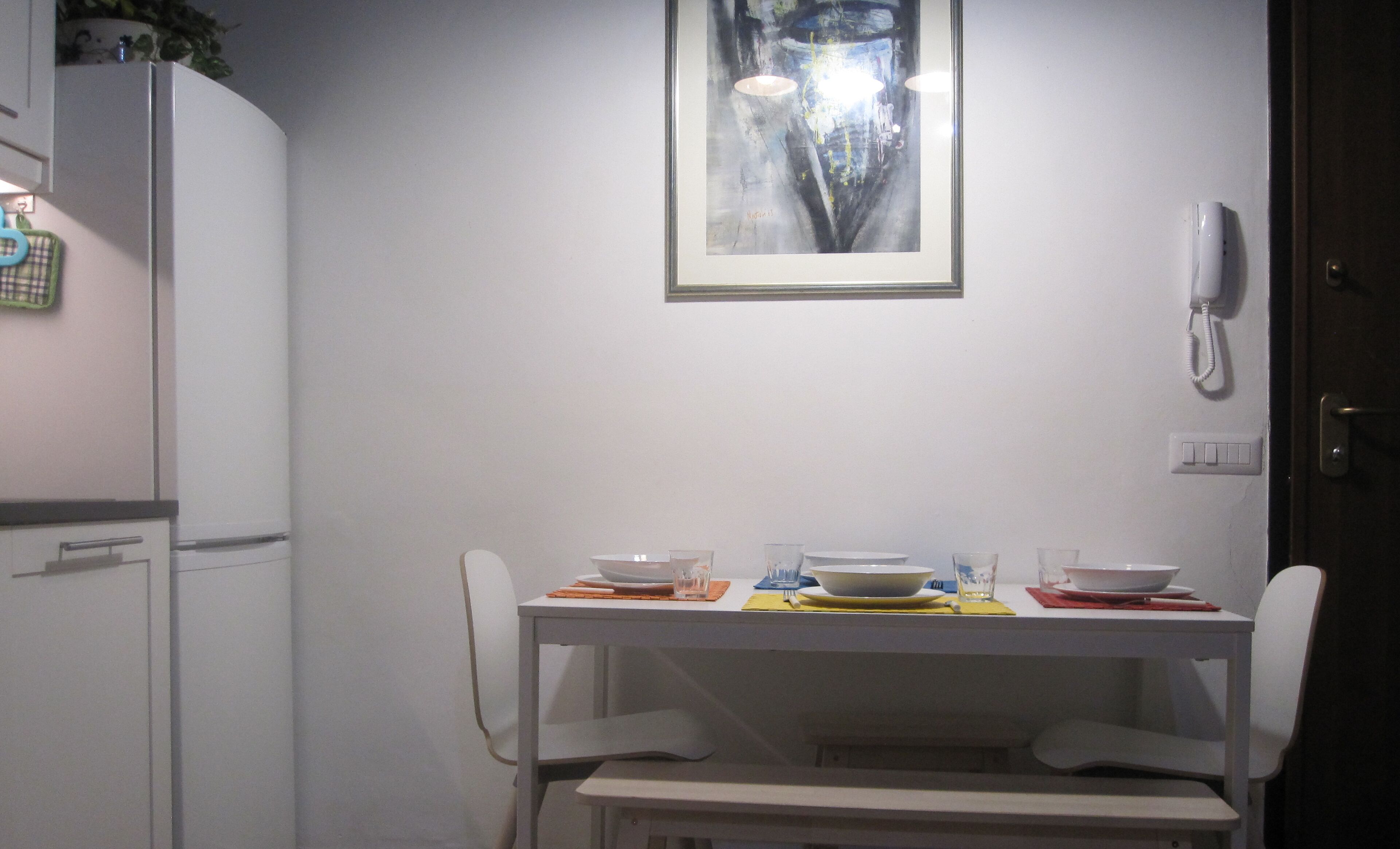 A dining area with a white table set for two, a framed abstract painting above the table, and a white telephone mounted on the wall to the right.