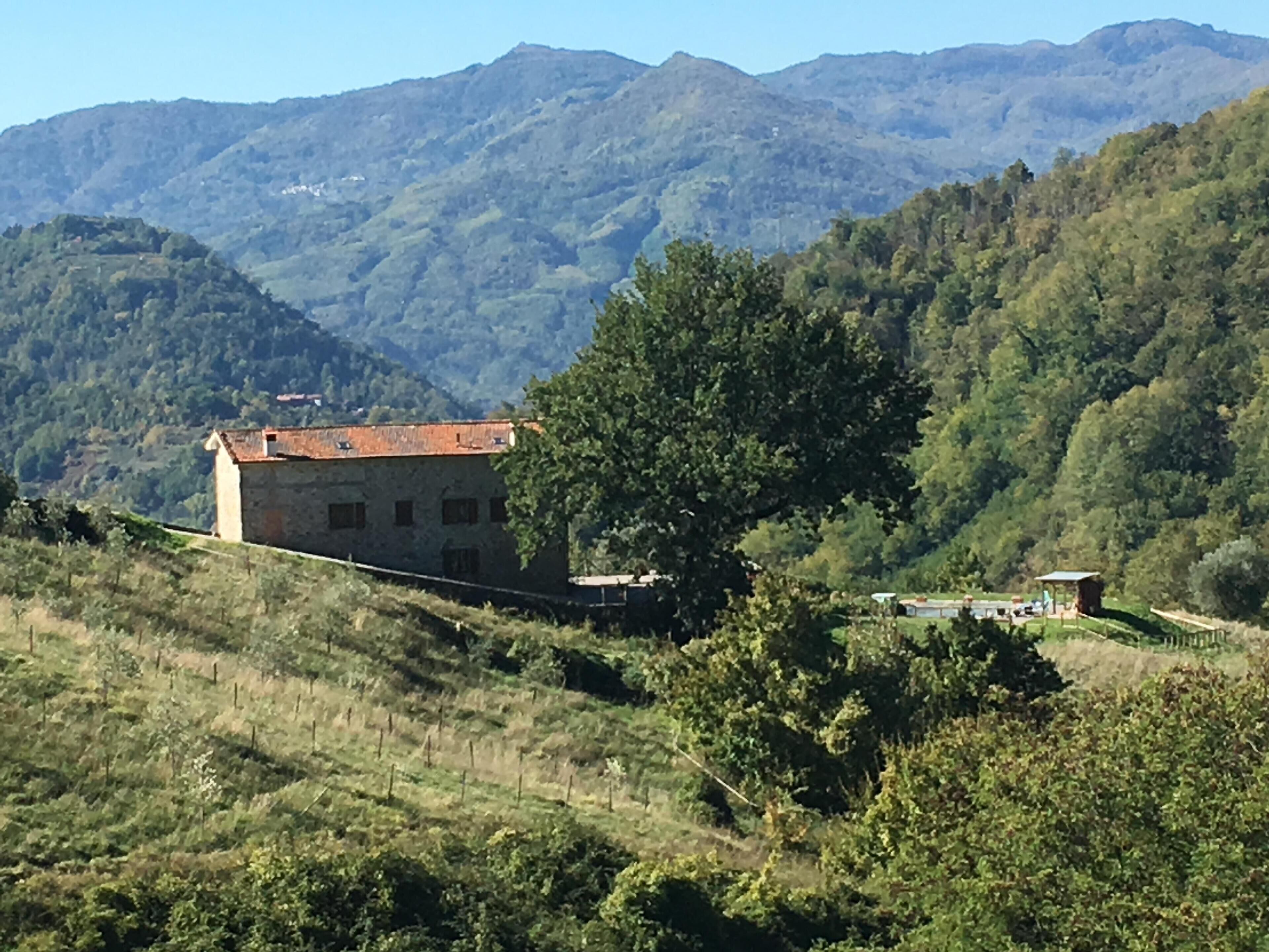 A rustic stone building with a red-tiled roof, surrounded by lush green hills and a clear blue sky.