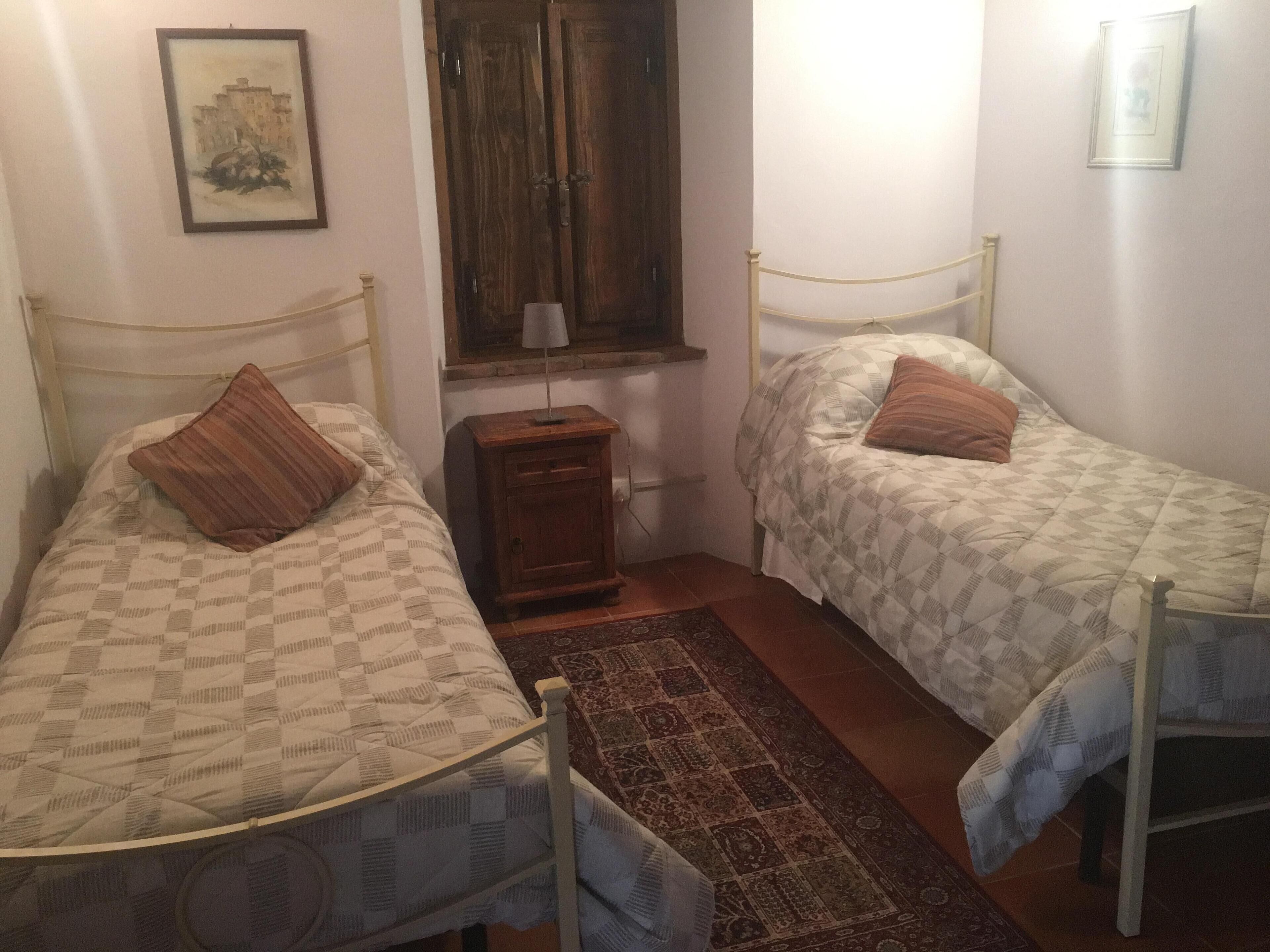 A room with two single beds, a small wooden nightstand, a window with a wooden shutter, and a framed picture on the wall.