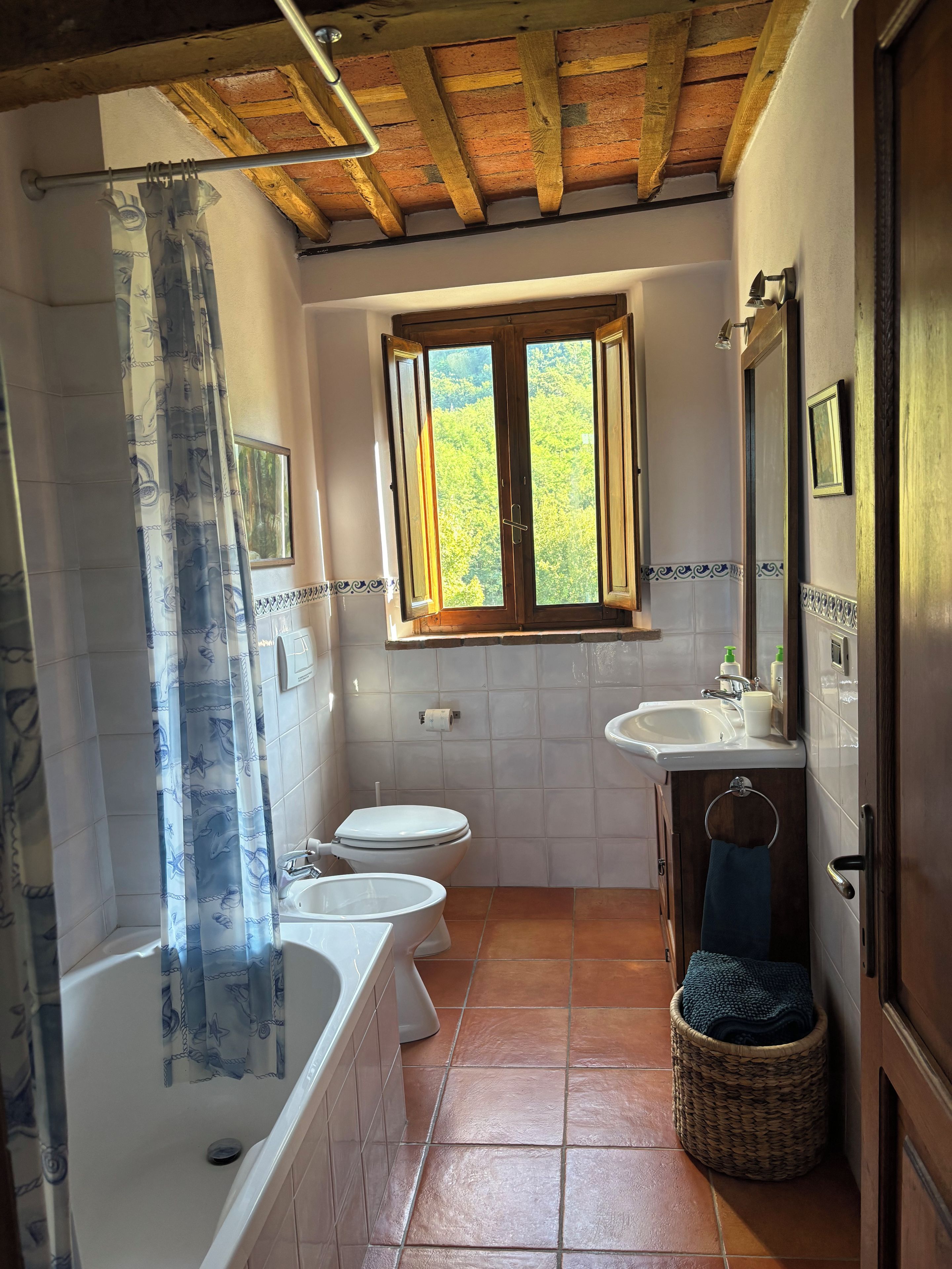 Ein Badezimmer mit Badewanne, Toilette und Waschbecken, ein Fenster mit Blick auf Grünflächen und eine Holzboden.
