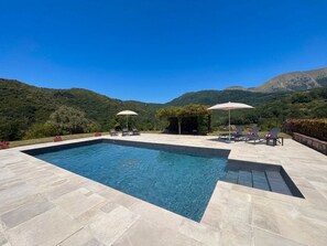 Piscina rectangular con solárium de baldosas, rodeada de sillas de descanso y sombrillas, con montañas al fondo.
