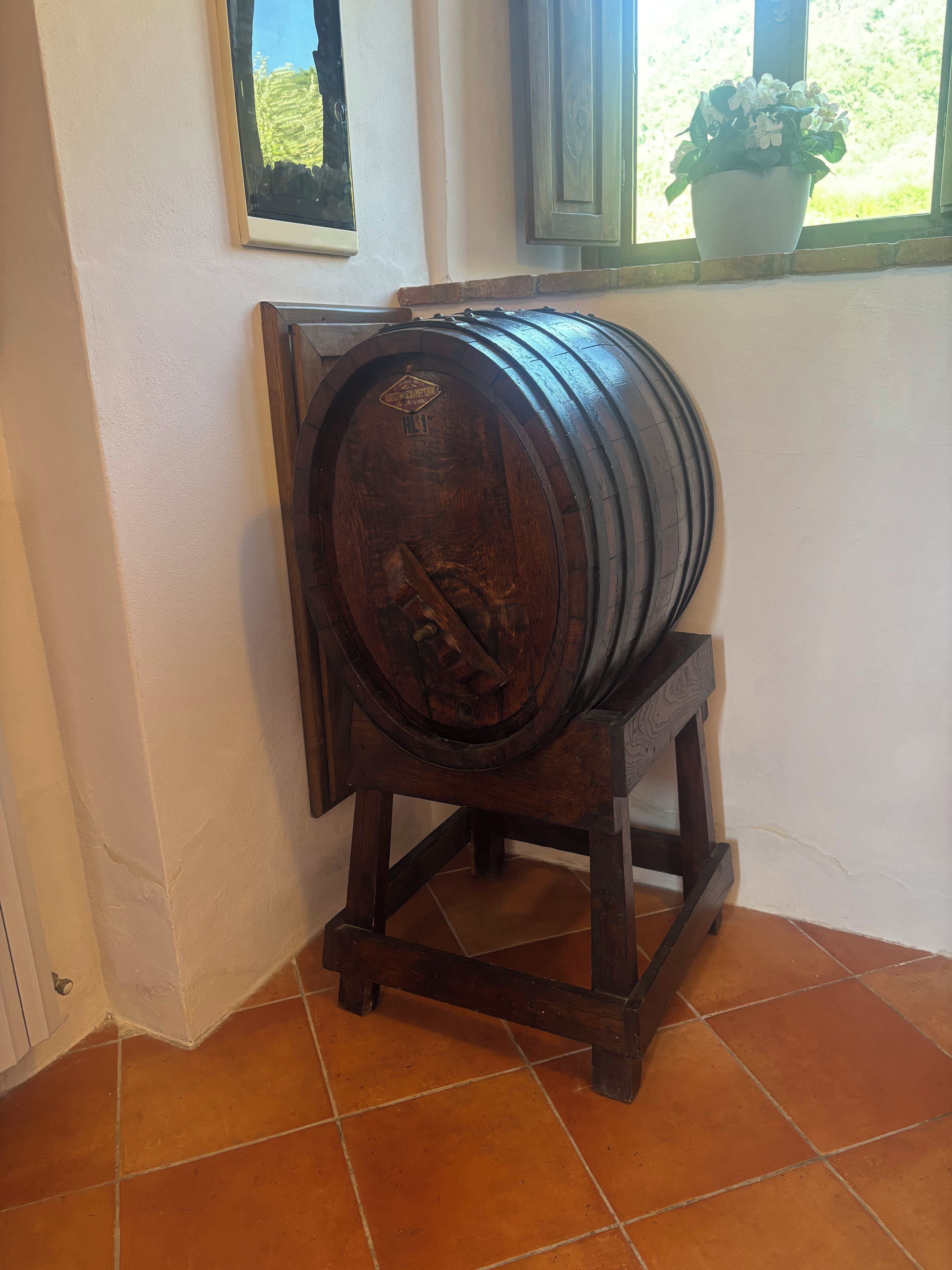 Un tonneau en bois, posé sur un support dans un coin, avec une fenêtre et une plante en pot en arrière-plan.