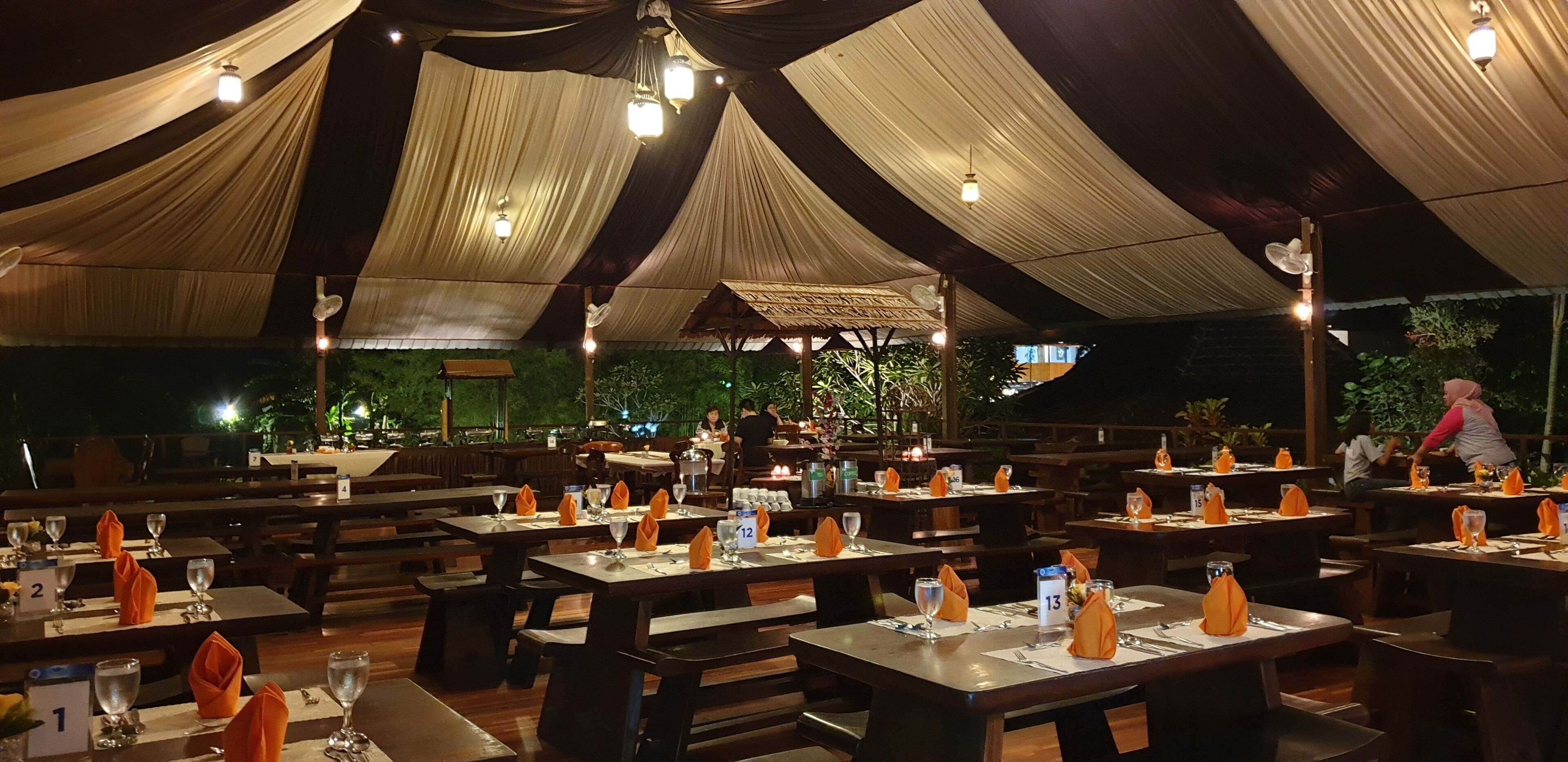 Restaurante al aire libre con mesas preparadas para comer, cubierto por una gran carpa con telas colgantes.