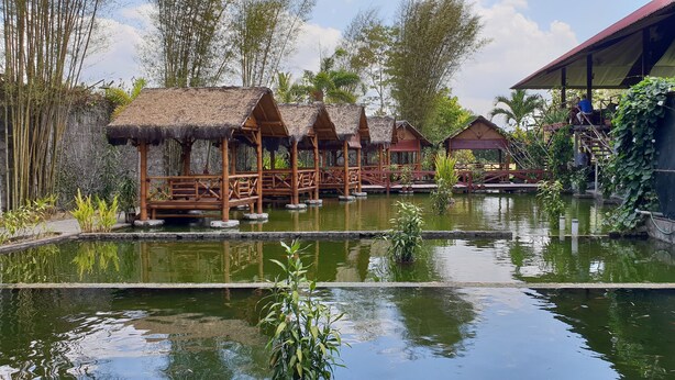 A series of wooden huts with thatched roofs over a reflective water body, surrounded by bamboo and tropical vegetation. - Borobudur Bed & Breakfast - Hostel (Mungkid)