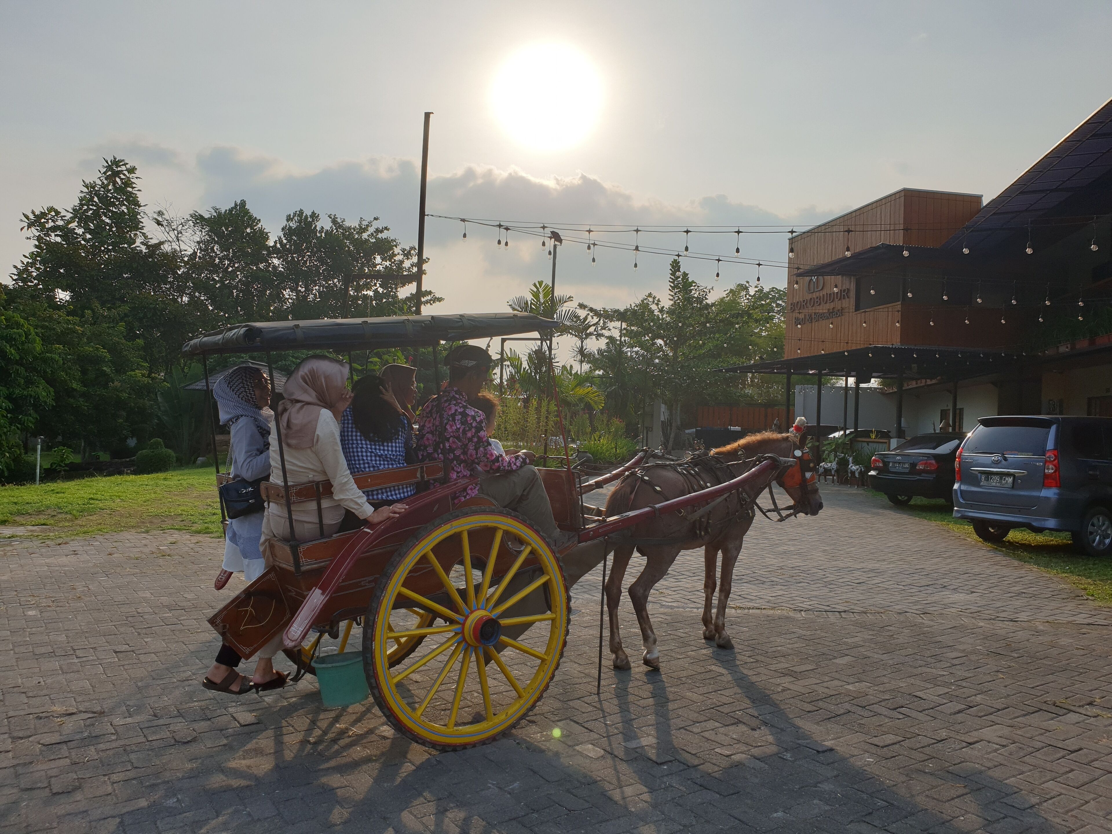 A horse-drawn carriage with passengers, parked on a paved area with buildings and greenery in the background.