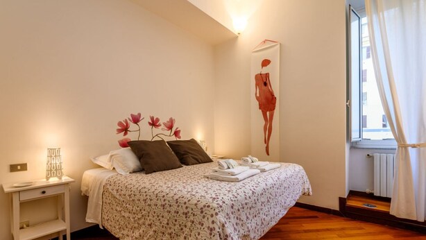 Un dormitorio con cama, mesita de noche, decoración en la pared y ventana con cortinas.