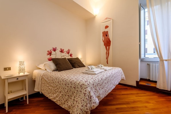 A bedroom with a bed, bedside table, wall art, and a window with curtains.