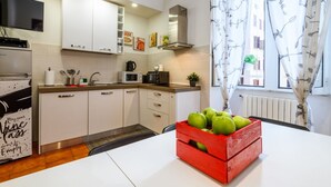 Una cocina moderna con armarios blancos, un microondas, un hervidor y una cesta roja con manzanas verdes sobre la mesa.