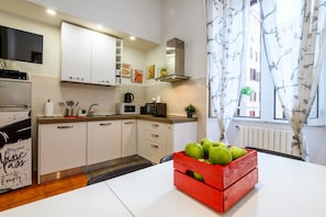 A modern kitchen with white cabinets, a microwave, a kettle, and a red crate of green apples on the table.