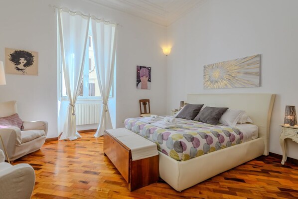 A bedroom with a bed, a chair, a bench, a window with curtains, and wall art.