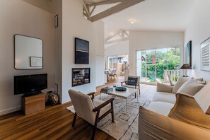 Sala de estar moderna com televisão de tela plana, piso de madeira, um sofá, uma mesa de centro e uma janela grande com vista para o exterior.