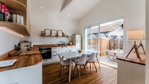 Une cuisine moderne avec des plans de travail en bois, un coin repas avec une table et des chaises blanches, et une grande porte-fenêtre coulissante donnant sur une terrasse extérieure.