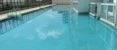 A clear blue pool with a tiled deck, surrounded by a black fence and a building with large windows.