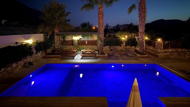 A swimming pool with blue lighting, surrounded by palm trees and stone walls, with outdoor seating and a pergola. - 5 Bedrooms, heated pool, pool table, table tennis, bbq & wifi. (Mugla)