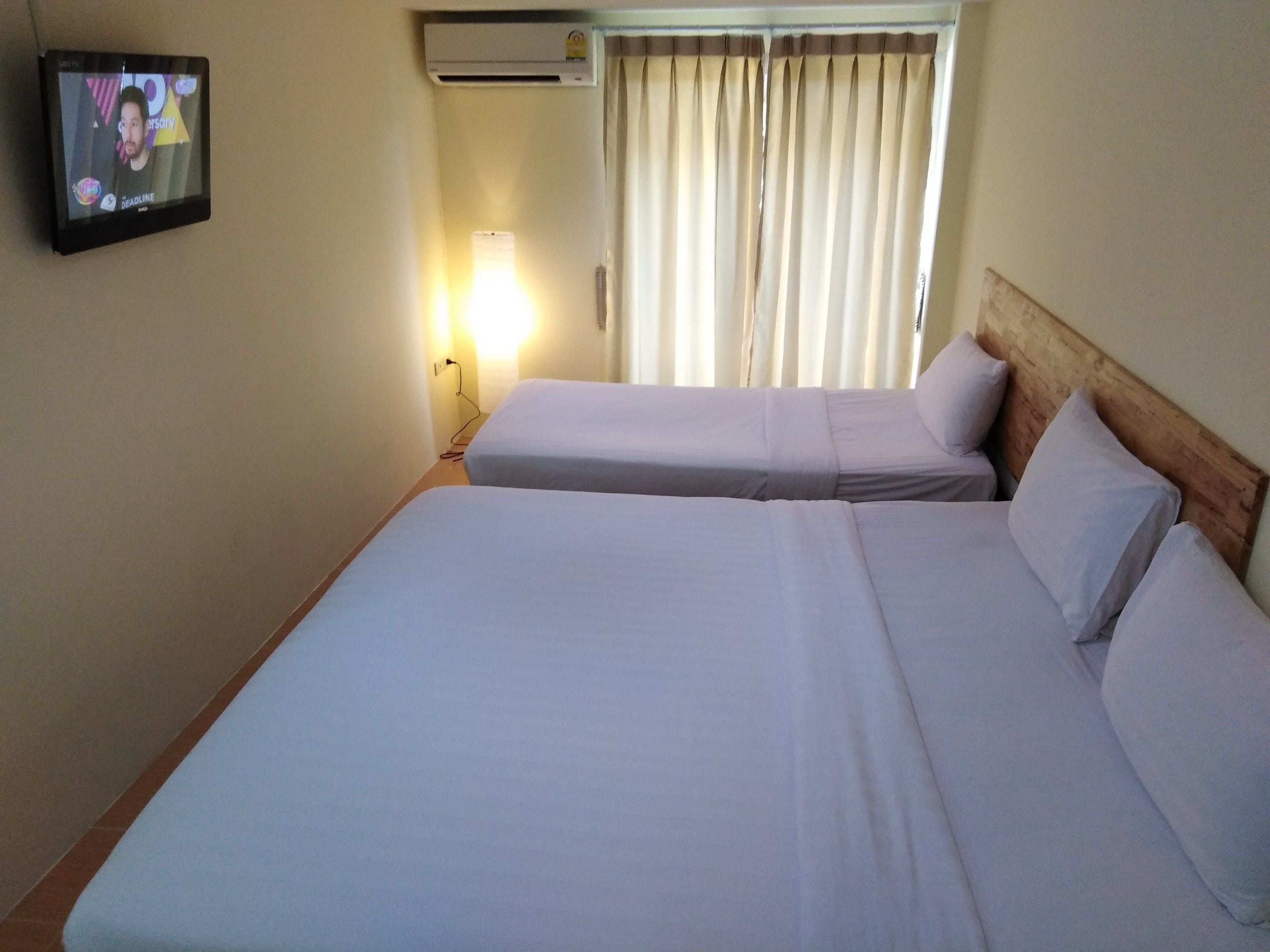 A hotel room with two beds, a TV on the wall, a window with curtains, and a wall-mounted air conditioner.