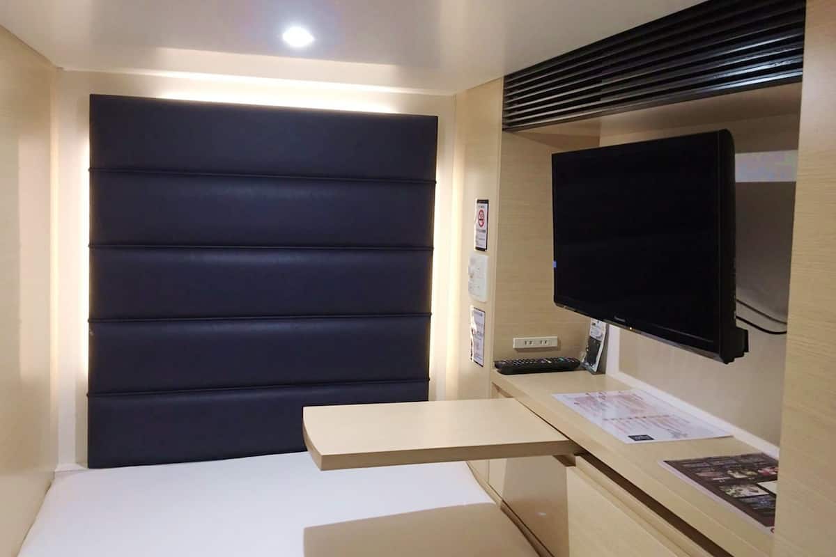 A hotel room with a large dark-colored headboard, a flat-screen TV mounted on the wall, a wooden desk with a flat surface, and a small shelf with papers.