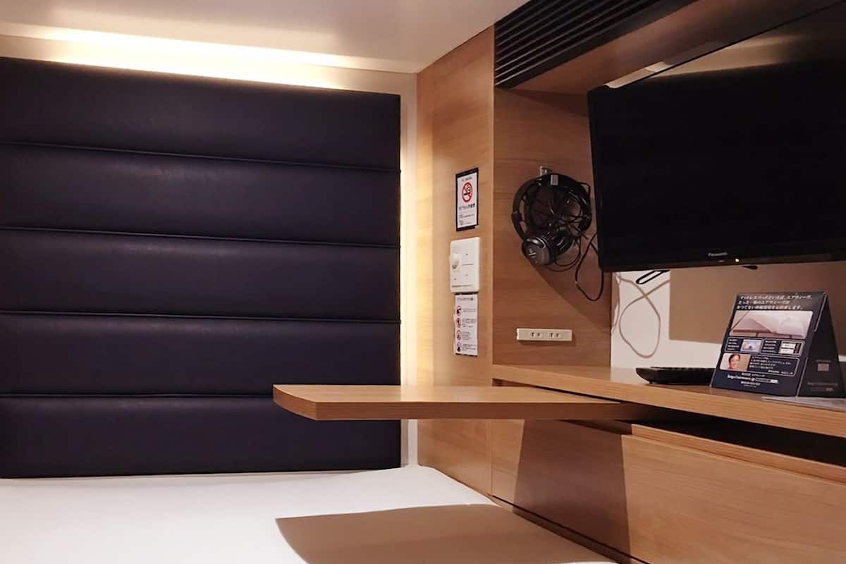 A modern hotel room with a dark padded headboard, a wooden bedside table, a flat-screen TV, and a wall-mounted sound system.