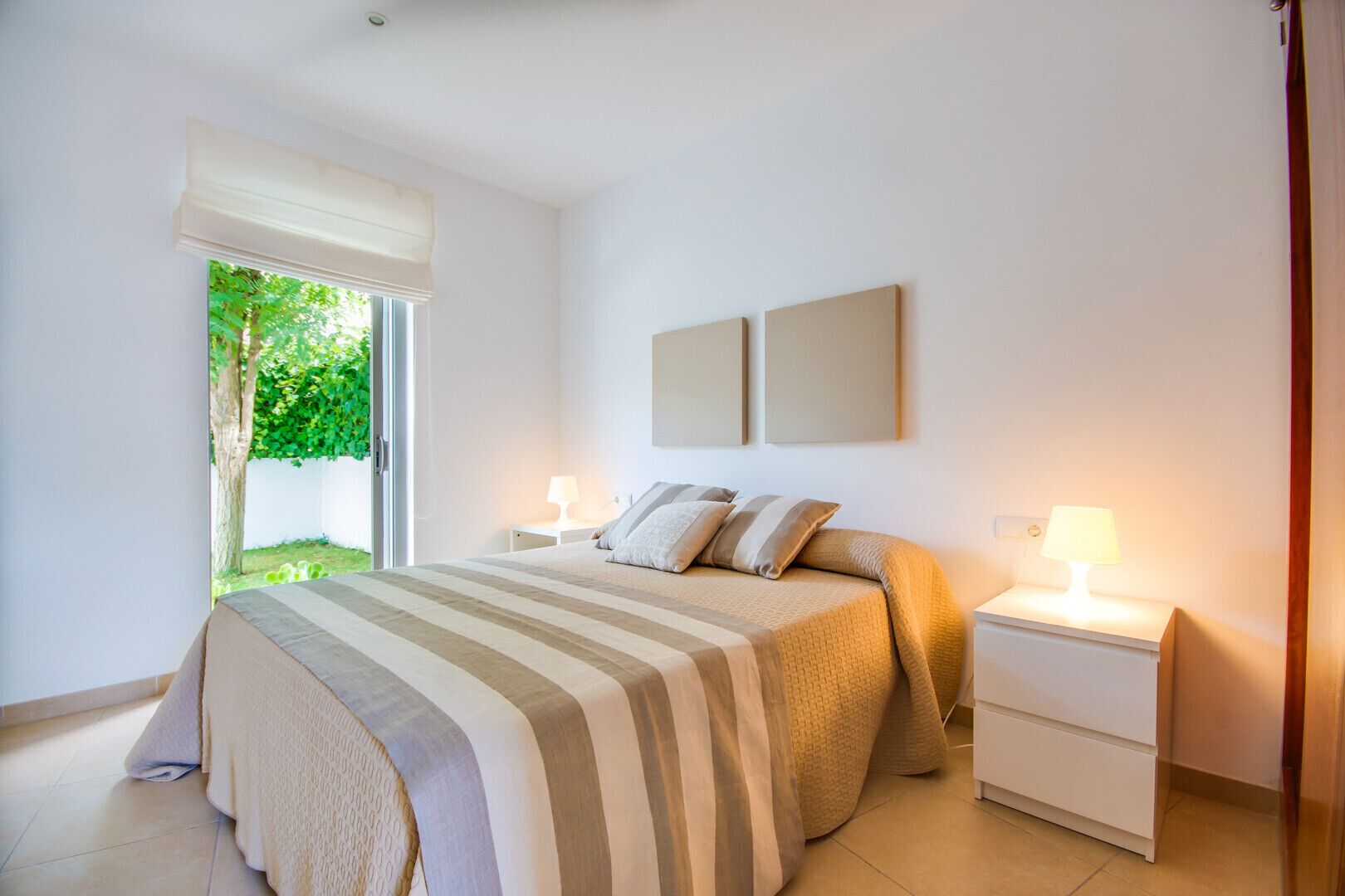 A bedroom with a large bed, two bedside tables with lamps, a window with a view of greenery, and a wall with two framed pictures.