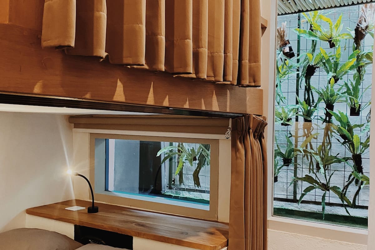 Un dormitorio con cama, mesita de noche, ventana con cortinas y vistas a plantas afuera.