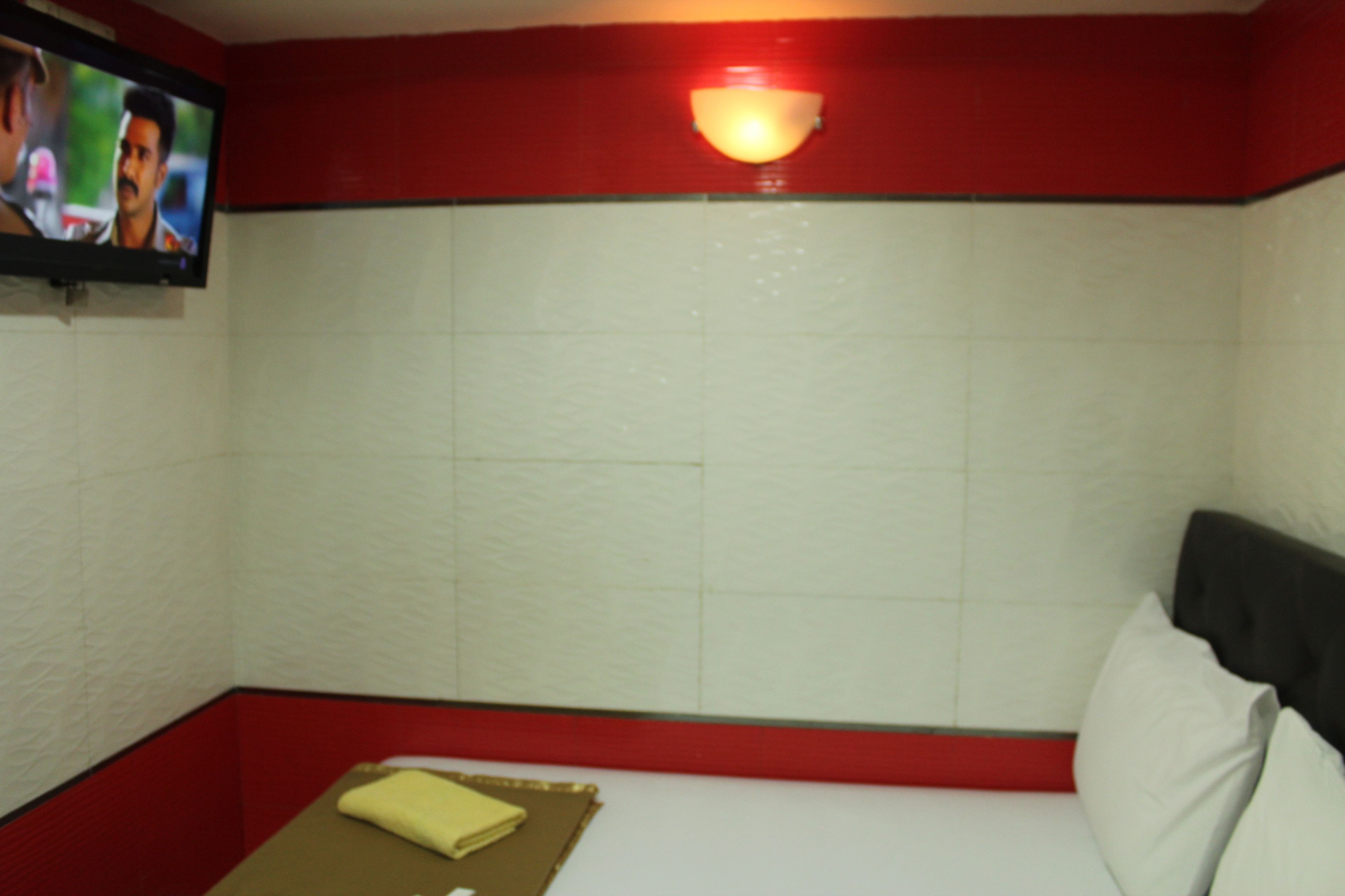 Quarto pequeno, com decoração em vermelho e branco, uma cama de solteiro, televisão na parede e luminária fixa.