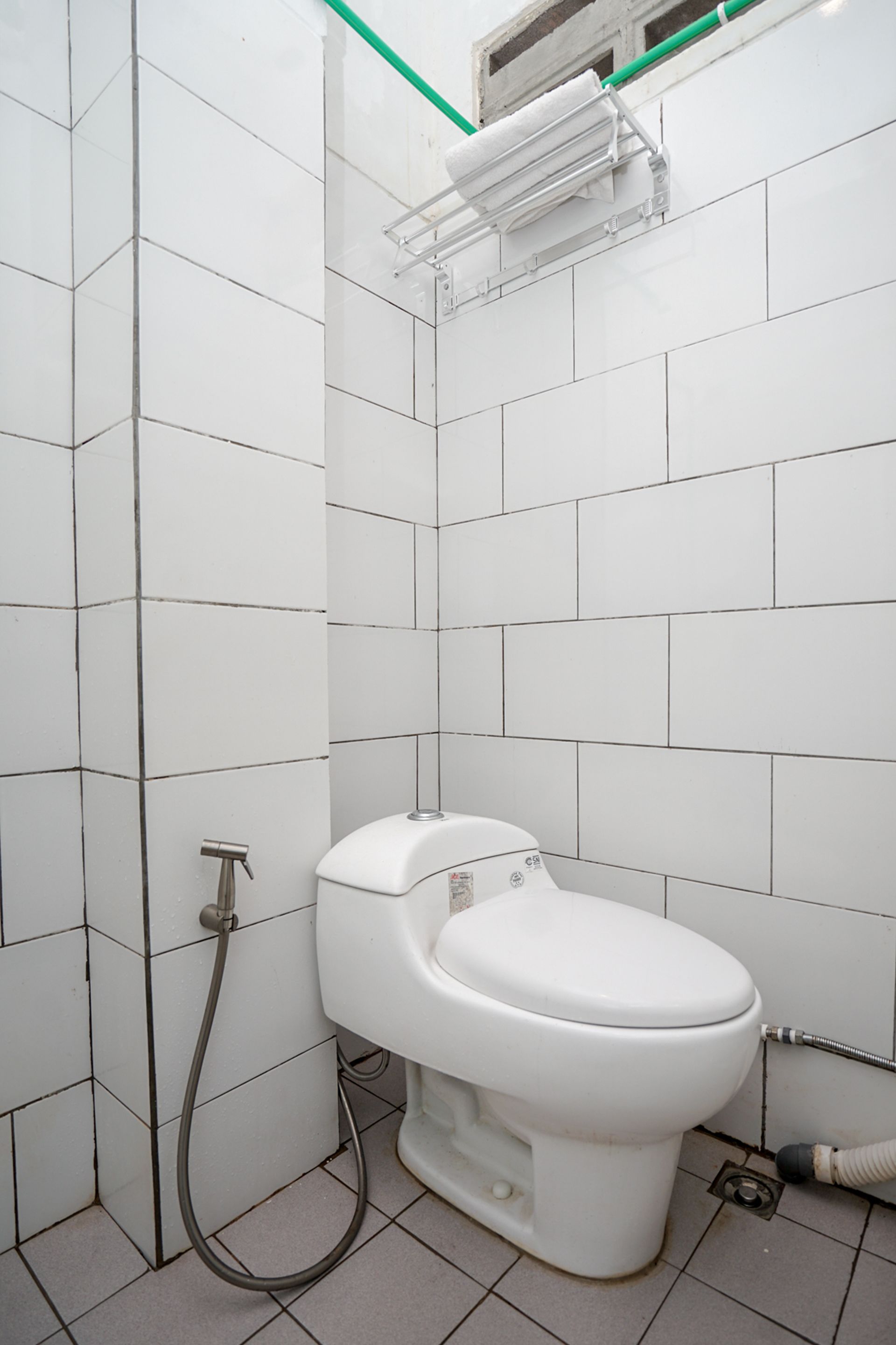 A small, white-tiled bathroom with a toilet, a shower hose, and a visible vent.