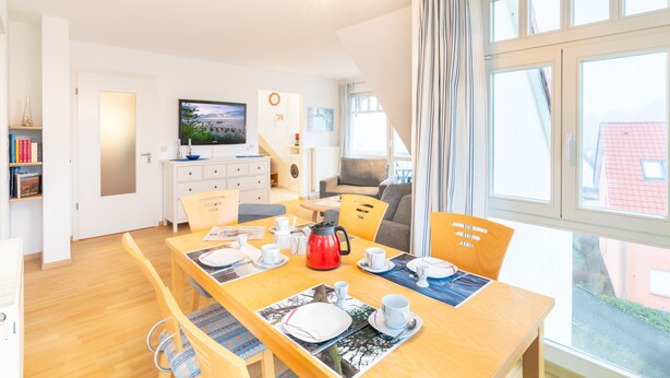 A bright, modern living room with a wooden dining table set for four, a red coffee pot, and a television on the wall. - Family-friendly vacation apartment Seeschwalbe in Koserow - Holiday apartment (Koserow)